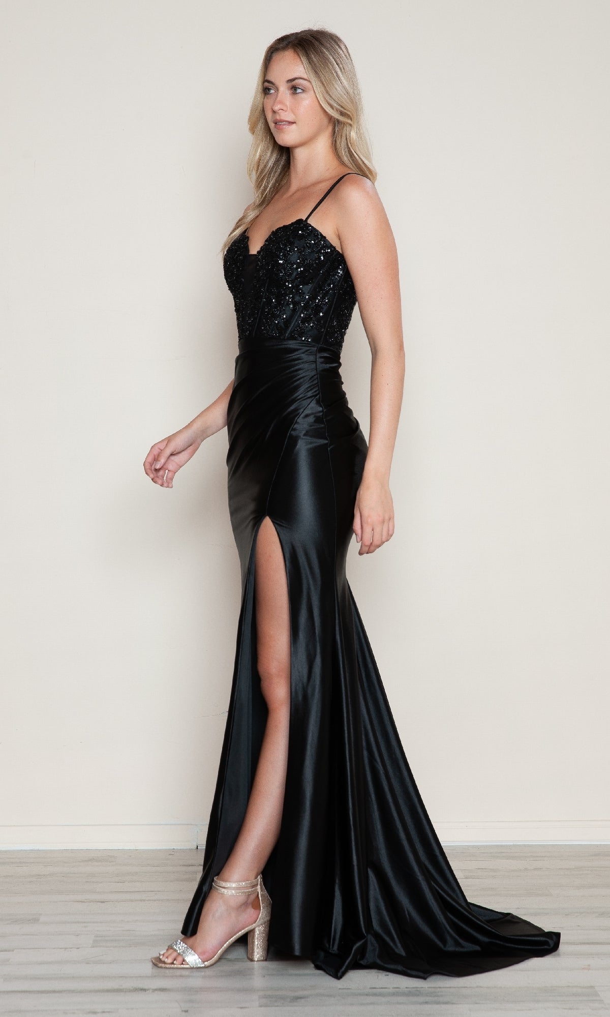 A woman in the Black Beaded-Bodice Long Prom Dress (Poly USA 9616), featuring thin straps, a beaded corset bodice, and a thigh-high slit, pairs it with high-heels as she stands against a plain light-colored wall.